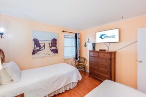 Ein Schlafzimmer mit Bett, Kommode, Stuhl und einem Bild von Strandstühlen. Ein Fernseher zeigt 'Big Fish Rentals'.