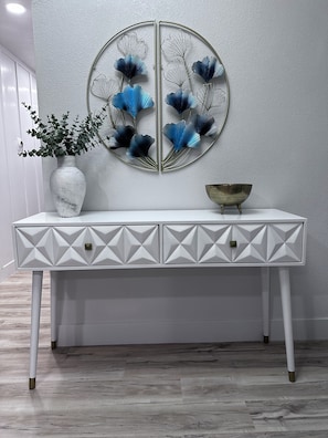 Une console blanche aux tiroirs à motifs géométriques, une œuvre d’art en métal à accrocher au mur, un vase orné de feuillage vert et un bol en métal.