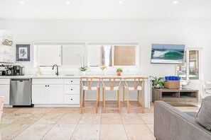 A modern kitchen with white cabinetry, a built-in dishwasher, and a central island with bar stools. - Charming 2BR Bungalow Steps from the Beach with Fire Pit (Newport Beach)