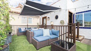 Un espacio de estar al aire libre con mobiliario de mimbre y una mesa de vidrio, rodeado por un muro blanco con detalles azules y un toldo negro.
