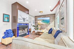 A living room with a fireplace, a large TV screen, a sofa, and a view of a beach. - Charming 3BR Bungalow Steps to Sand with Deck and Fire Pit (Newport Beach)