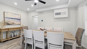 Un comedor con una mesa de madera, sillas blancas, un ventilador de techo y un equipo de aire acondicionado fijado a la pared.