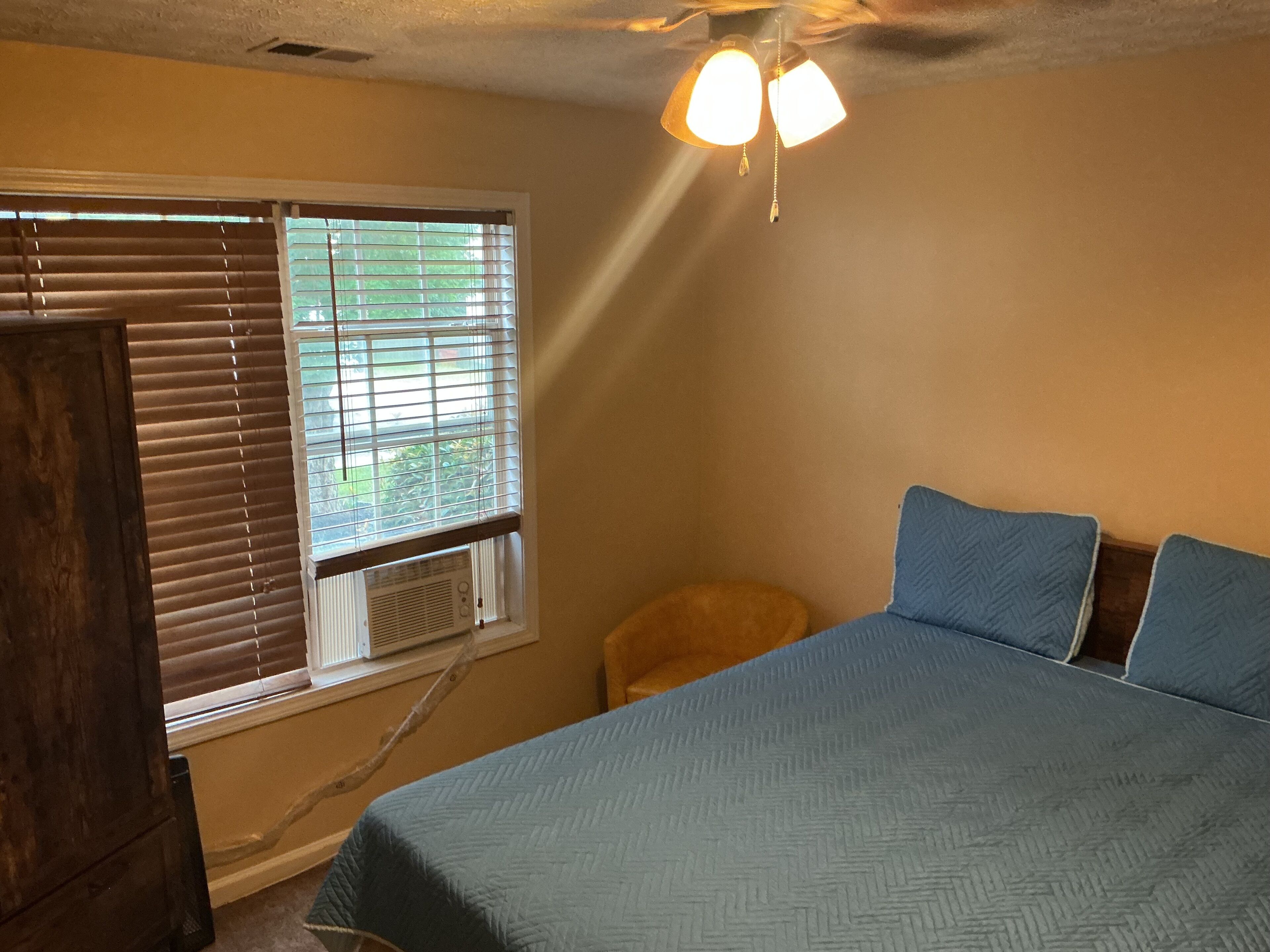 A bedroom with a bed, a window with blinds, a ceiling fan, and a small stool.