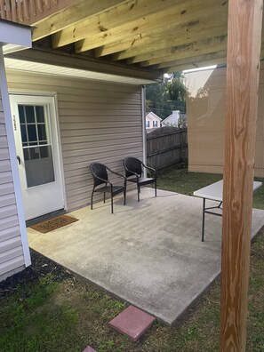 A covered patio area with two chairs and a table, a door with a window, and a wooden fence in the background. - Enchanting Guest Studio with backyard vibe (Conley)