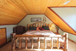 A bedroom with a sloped ceiling, a wooden bed frame, a nightstand, a lamp, and a chair. - The Lodge at Stone Pile Gap (Bryson City)