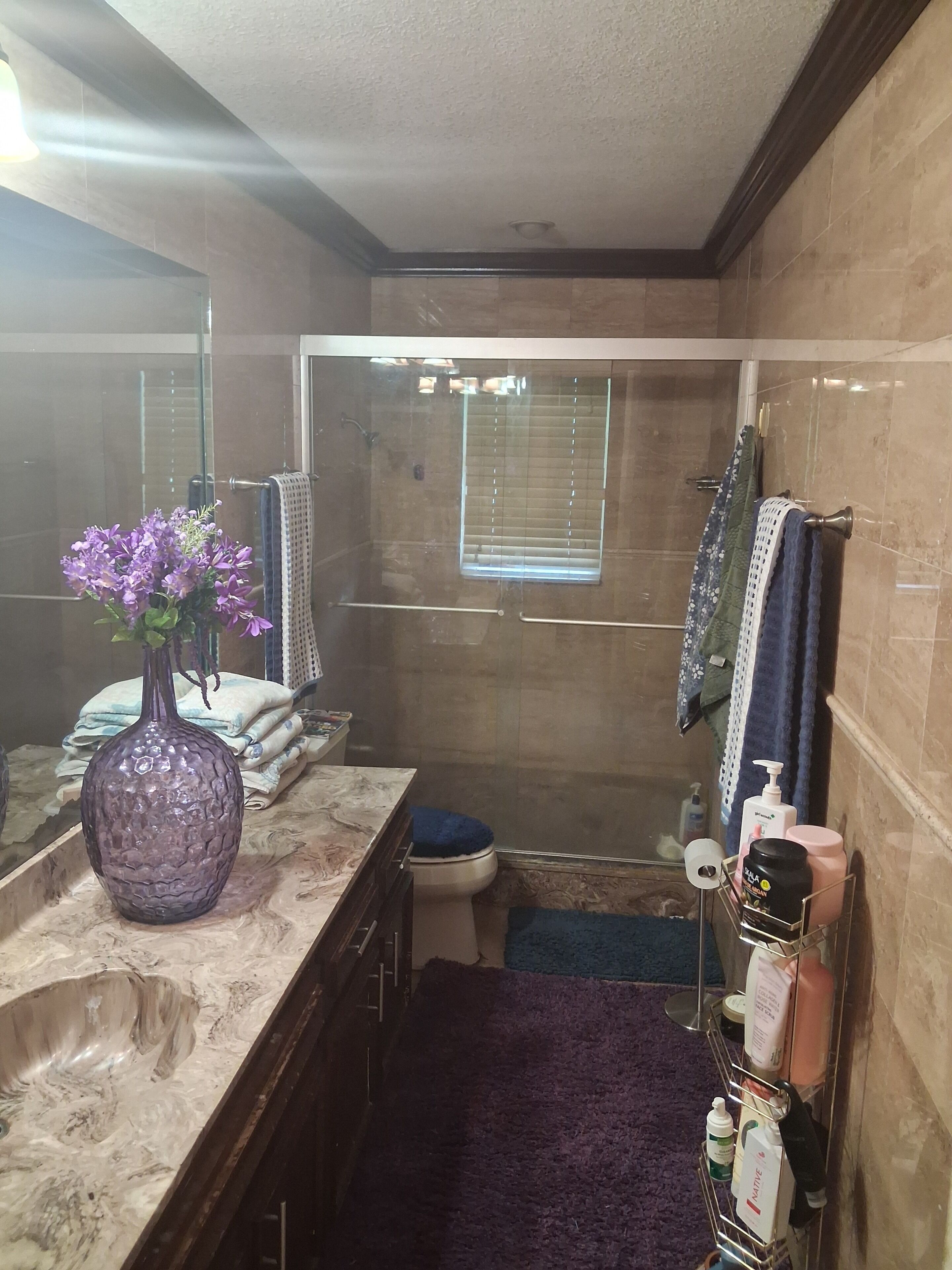 A bathroom with a marble countertop, a purple vase, a shower with a glass door, and a towel rack with multiple towels.