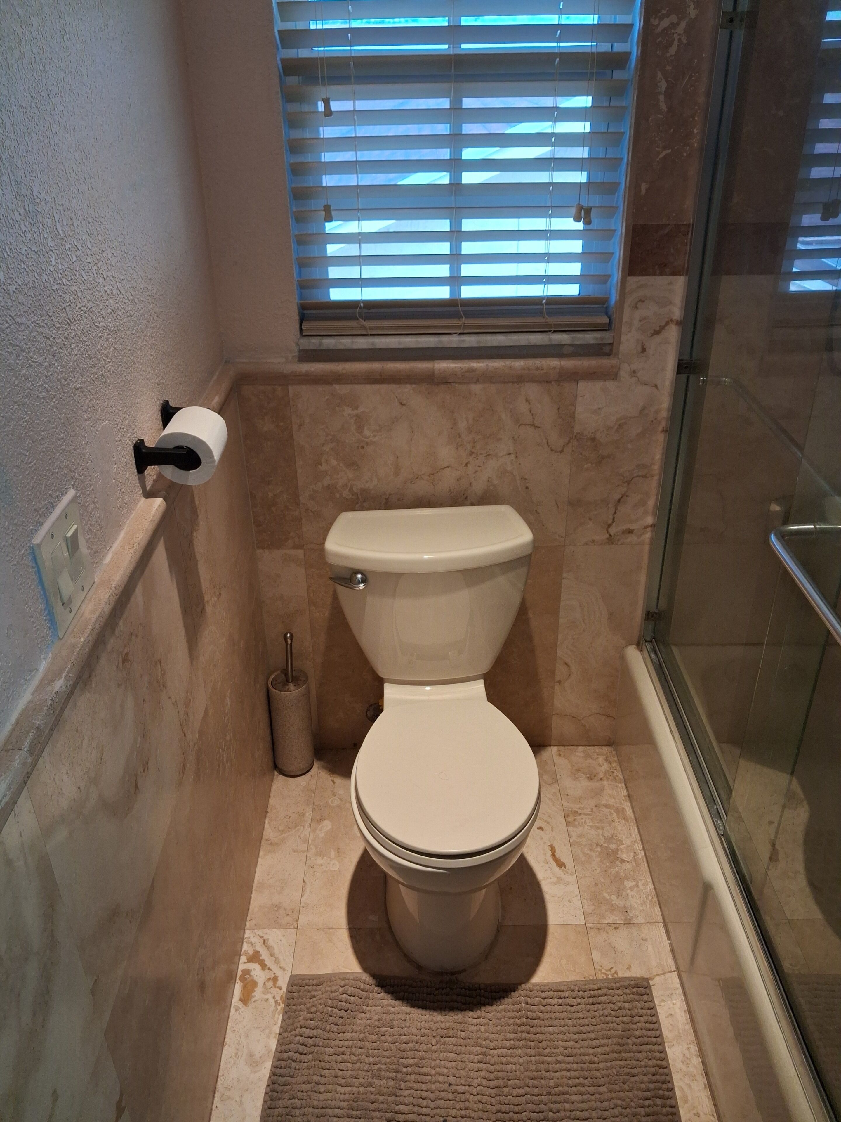 A bathroom with a toilet, a window with blinds, a shower area, and a marble floor.