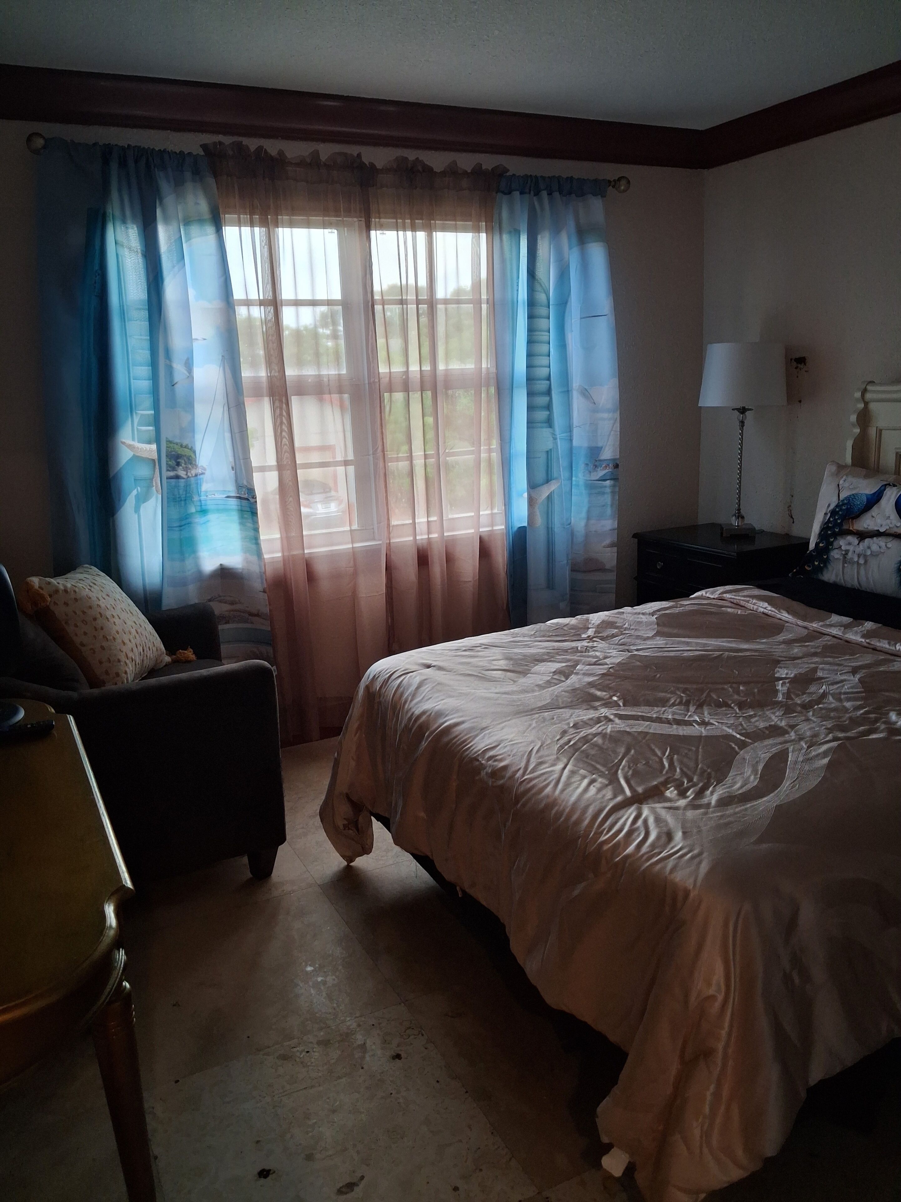 A bedroom with a bed, a chair, a nightstand with a lamp, and a window with curtains.