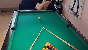 A pool table with a cue stick and balls, a sofa, and a bookshelf in a room with a window.