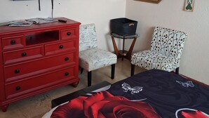 Um quarto com cama, penteadeira vermelha, televisão, duas cadeiras, uma mesinha e decorações nas paredes.