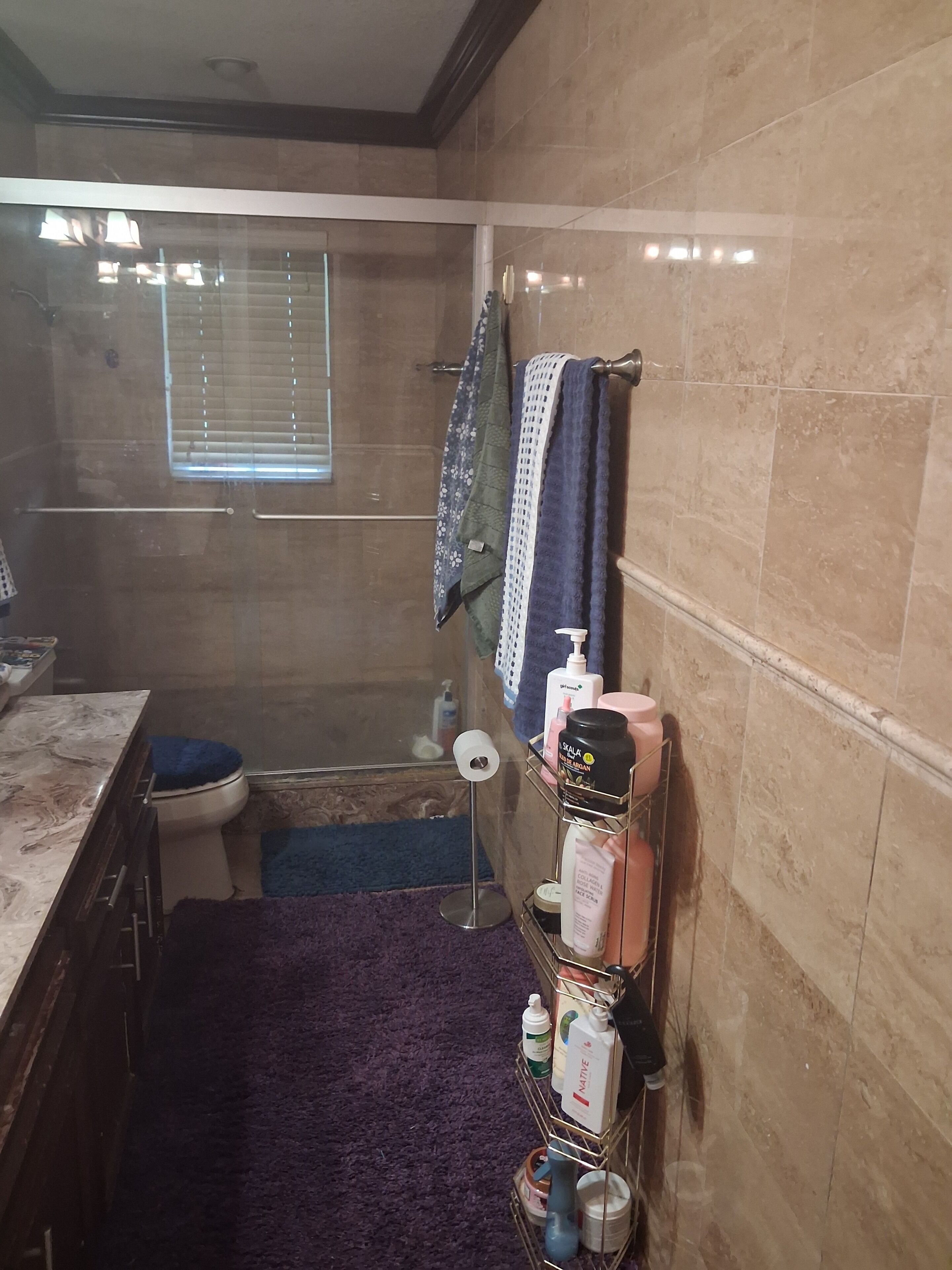 A bathroom with a toilet, a shower, and a sink area. There are towels hanging on the wall, a shelf with toiletries, and a window with blinds.