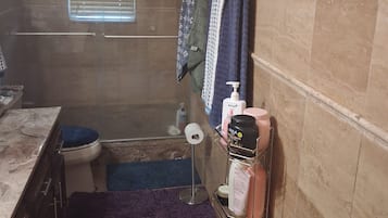 A bathroom with a toilet, a shower, and a sink area. There are towels hanging on the wall, a shelf with toiletries, and a window with blinds.