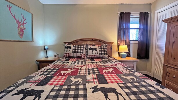 A bedroom with a bed featuring a checkered quilt with animal prints, a wooden headboard, a nightstand with a lamp, a window with curtains, and a wall-mounted artwork. - On Top of the World- Amazing Views (Bryson City)