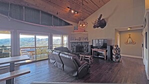 A spacious living room with a fireplace, a mounted moose head, and a large window overlooking a mountainous landscape. - On Top of the World- Amazing Views (Bryson City)
