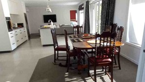 Un coin repas avec une table et des chaises en bois, une cuisine à l’arrière-plan et une grande fenêtre avec des rideaux.