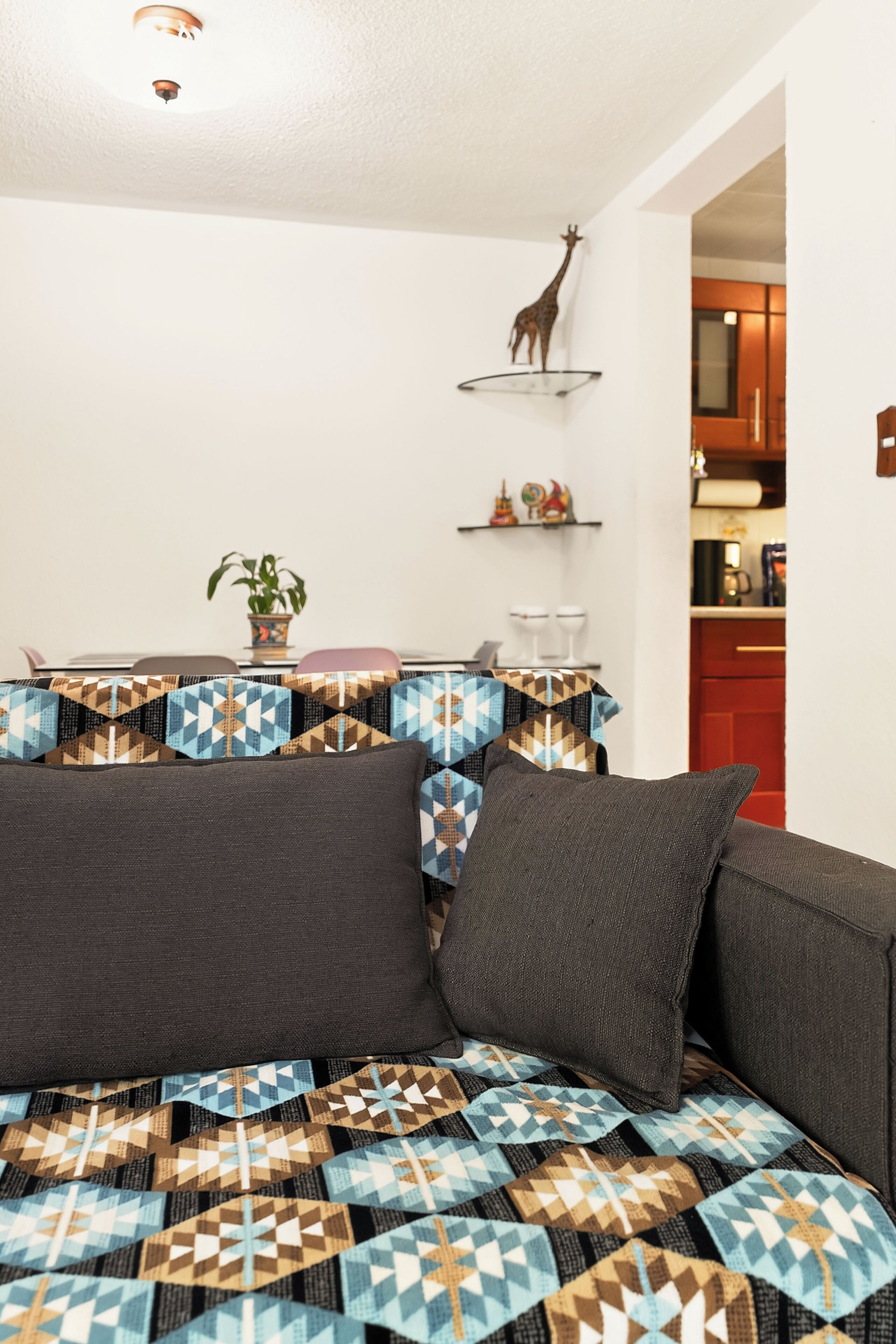 Una habitación con una cama, una planta en maceta, un estante con adornos y una cocina al fondo.