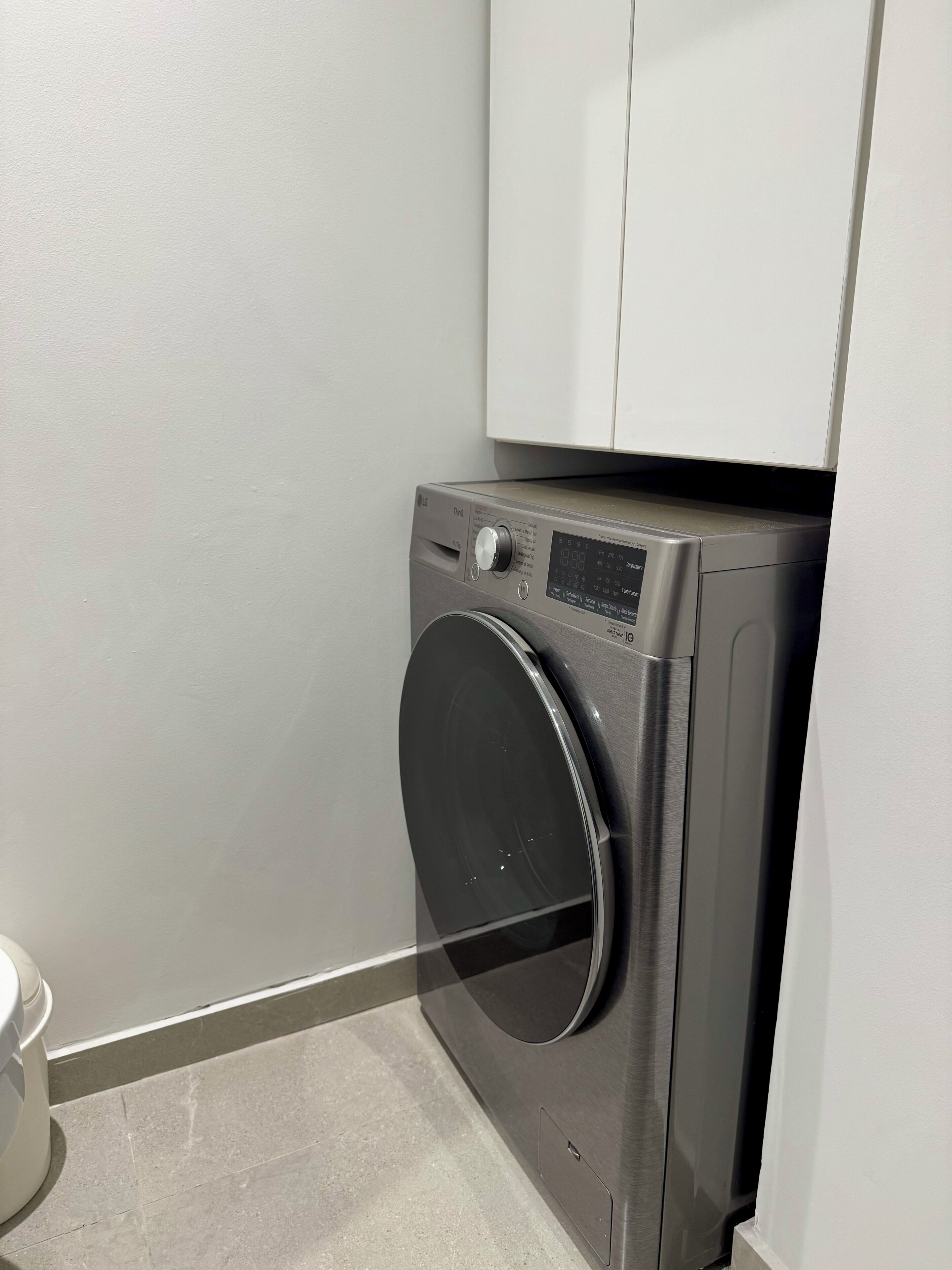 A compact, modern washing machine with a control panel, situated in a corner of a room with a plain wall and a tiled floor.