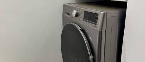 A compact, modern washing machine with a control panel, situated in a corner of a room with a plain wall and a tiled floor.
