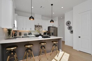 A modern kitchen with white cabinets, a central island with stools, pendant lighting, and decorative wall art. - Midtown Home – Free Parking, W/D, Private Yard (Houston)