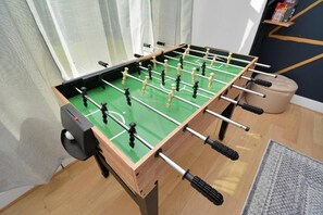 A foosball table in a room with a wooden floor, a window with curtains, and a small shelf with items. - Pet-Friendly Midtown House, Private Yard & BBQ (Houston)