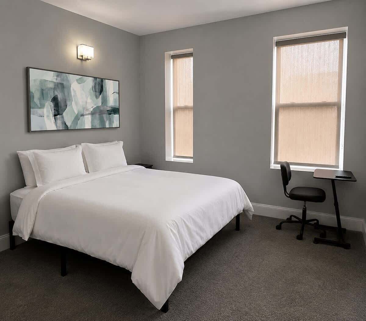 A bedroom with a bed, a desk, two windows, and a wall-mounted light fixture.