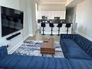 Ein modernes Wohnzimmer mit einem großen Flachbildfernseher, einem blauen Sofa, einem Holztisch und einer Küchenzeile mit weißen Schränken und einem Barhocker.