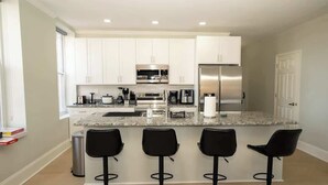 A modern kitchen with white cabinets, stainless steel appliances, a granite countertop, and three black bar stools. - Spacious PennStation Downtown Penthouse W/ Parking (Baltimore)
