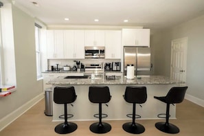 A modern kitchen with white cabinets, stainless steel appliances, a granite countertop, and three black bar stools. - Spacious PennStation Downtown Penthouse W/ Parking (Baltimore)