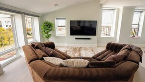 A modern living room with a large brown sectional sofa, a flat-screen TV mounted on the wall, and large windows providing a view of the outdoors. - Spacious PennStation Downtown Penthouse W/ Parking (Baltimore)