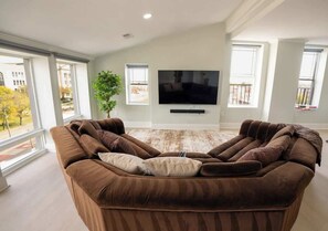 A modern living room with a large brown sectional sofa, a flat-screen TV mounted on the wall, and large windows providing a view of the outdoors. - Spacious PennStation Downtown Penthouse W/ Parking (Baltimore)
