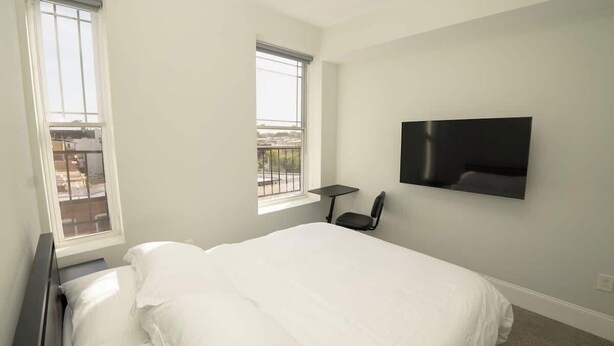 A bedroom with a bed, two windows, a TV mounted on the wall, and a small table. - Massive Downtown Penn Station apt with parking (Baltimore)