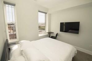 A bedroom with a bed, two windows, a TV mounted on the wall, and a small table. - Spacious Arts District Apt w/ Free Parking, Pet (Baltimore)