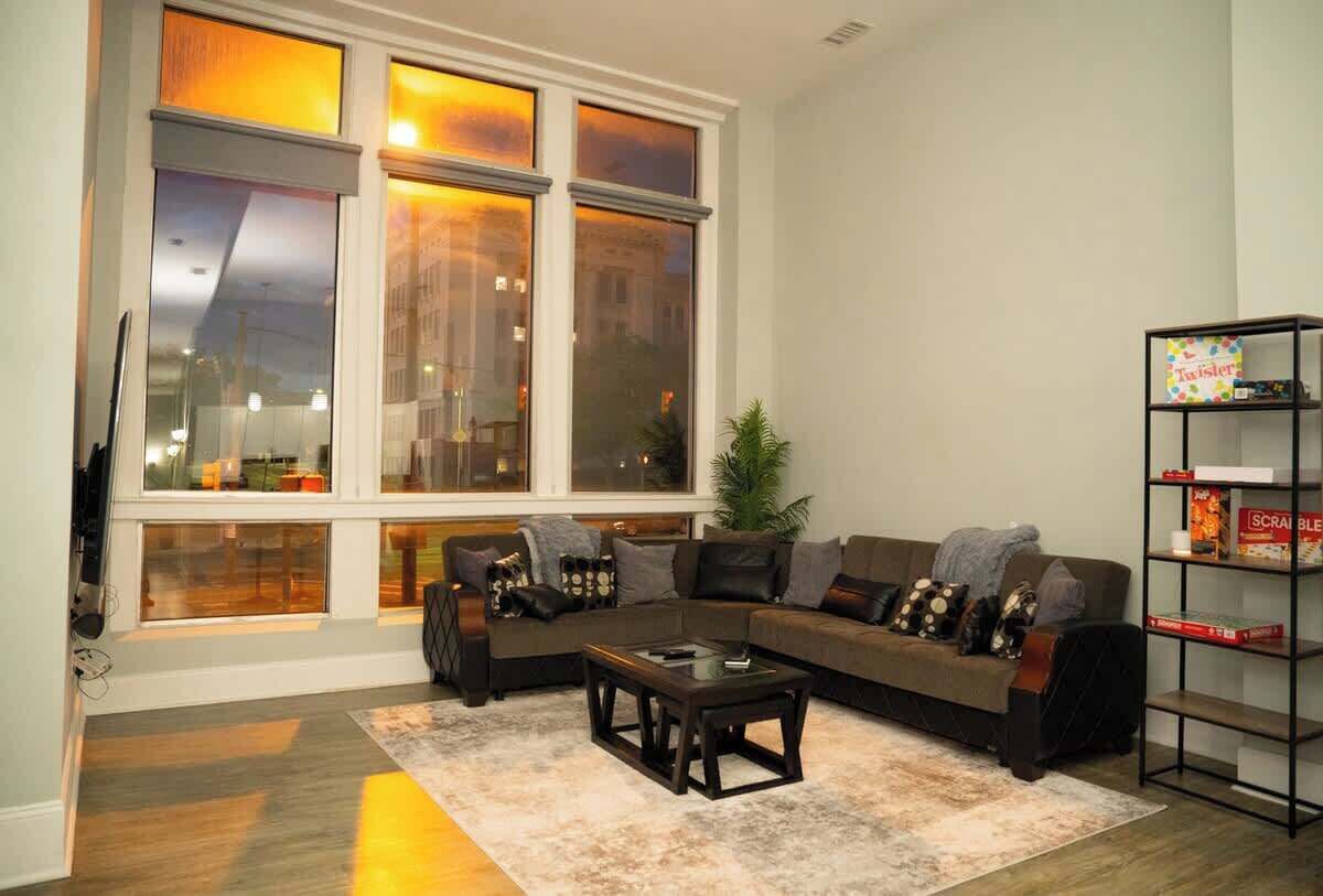 A living room with a sectional sofa, a coffee table, a bookshelf, and large windows.