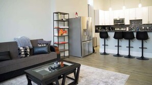 A modern living room with a sofa, coffee table, and a kitchen area with a refrigerator, microwave, and bar stools. - Massive Downtown Penn Station apt with parking (Baltimore)