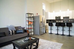 A modern living room with a sofa, coffee table, and a kitchen area with a refrigerator, microwave, and bar stools. - Massive Downtown Penn Station apt with parking (Baltimore)