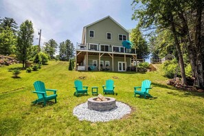 A two-story house with a porch, a fire pit, and four green Adirondack chairs. - Serenity Point- Shellcamp Lakefront- 4bd3.5bth-Mountain Views- Pet Friendly! (Gilmanton)