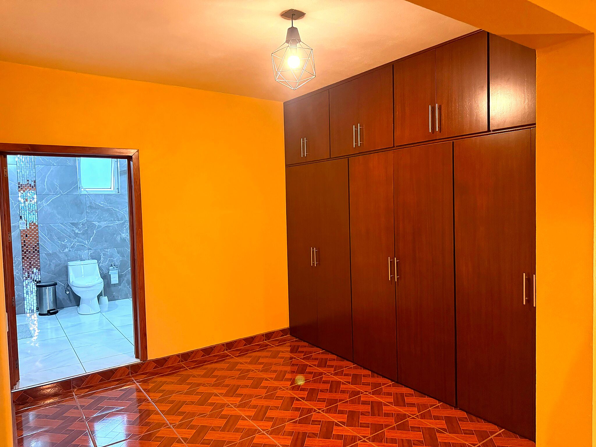 A room with a wooden wardrobe, a tiled floor, and a bathroom visible through a glass door.