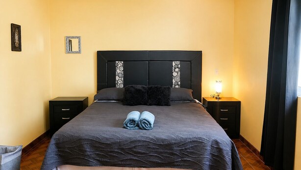 A neatly made bed with a dark patterned bedspread, two pillows, and rolled towels on the bed. There are two bedside tables with lamps, a small trash bin, and a wall-mounted mirror.