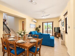 A dining area with a wooden table set for four, a blue sofa, and a television in the background. - Villa Saint Nicholas by Ezoria Villas (Agia Thekla)