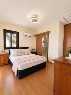 A bedroom with a bed, bedside tables, a wardrobe, a window with blinds, an air conditioning unit, and a ceiling light fixture. - Villa Saint Nicholas by Ezoria Villas (Agia Thekla)