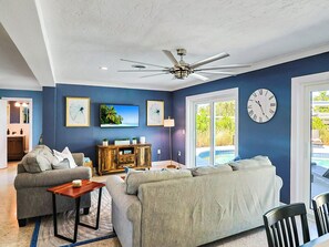 A living room with a ceiling fan, a large clock, and a television on the wall. - Siesta Key 3BR w/ Heated Pool, Game Room & Lanai (Sarasota)