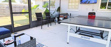 Une terrasse extérieure équipée d’une table de ping-pong, de chaises, d’une table en osier et d’un téléviseur fixé au mur.