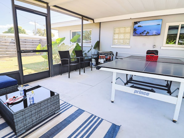 An outdoor patio with a ping pong table, chairs, a wicker table, and a TV mounted on the wall.