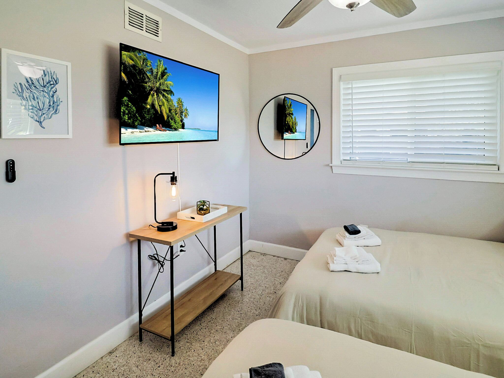 A bedroom with a bed, a desk with a lamp, a TV on the wall, and a window with blinds.