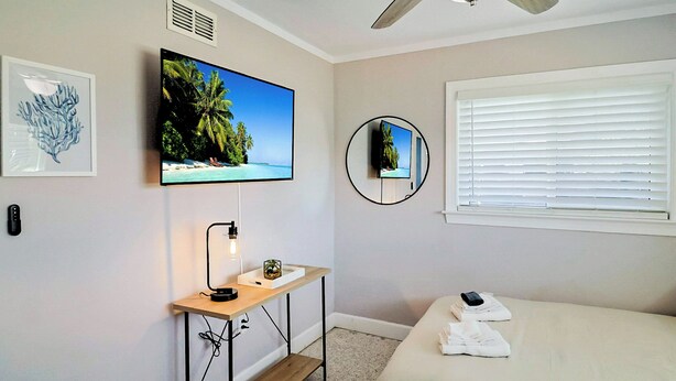 Une chambre à coucher avec un lit, un bureau avec une lampe, un téléviseur fixé au mur et une fenêtre avec des stores.