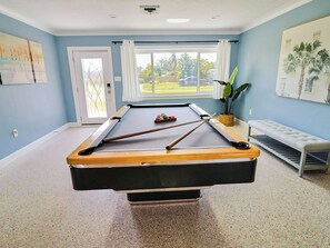 A pool table with cues and balls, a bench, a potted plant, and a window with a view of the outdoors.