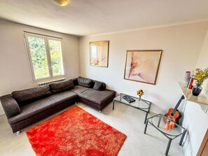Ein modernes Wohnzimmer mit einem Ecksofa, einem roten Teppich, einem Glastisch und einer Gitarre.