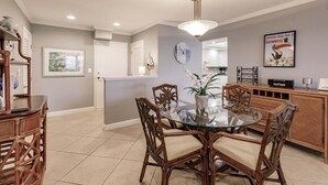 A dining area with a glass table and wooden chairs, a kitchen in the background, and a wall clock. - 1200-406 - Standard Plus - Embrace Spectacular Water Views (Siesta Key)