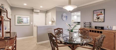 A dining area with a glass table and wooden chairs, a kitchen in the background, and a wall clock.
