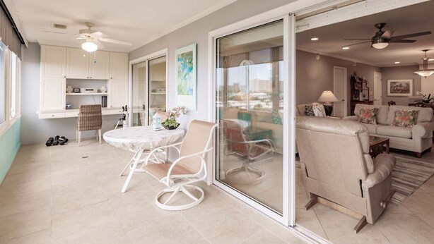 A spacious living area with a sliding glass door leading to a balcony, a dining area with a round table and chairs, and a kitchen with white cabinets. - 1200-406 - Standard Plus - Embrace Spectacular Water Views (Siesta Key)